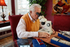 Elderly numismatist looking at coins through a microscope in his home office.