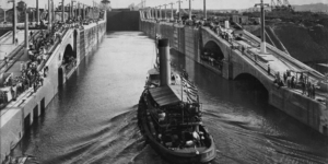 Black and white photograph of a steam-powered vessel navigating through the Miraflores locks of the Panama Canal surrounded by crowds of spectators lining the lock walls.